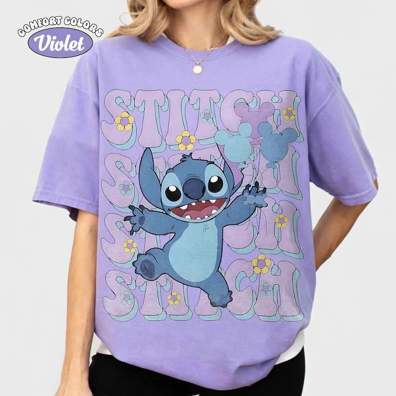 May include: A violet short-sleeve t-shirt featuring a graphic of the blue character Stitch holding light blue balloons. The background includes repeating text that reads STITCH in a wavy font, accented with small yellow and light blue flowers.