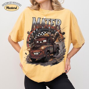 May include: Mustard-colored t-shirt featuring a graphic of Mater from the movie Cars. The design includes the name "MATER" in bold letters above the character, with a tire and lightning bolt background. The shirt has the words "Comfort Colors" and "Mustard" in a circle.