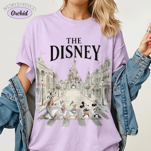 May include: Orchid Comfort Colors t-shirt with "THE DISNEY" printed in black. The shirt features a graphic of Disney characters crossing a crosswalk in front of a castle and buildings. The shirt is light purple.
