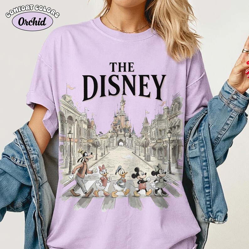 May include: Orchid Comfort Colors t-shirt with "THE DISNEY" printed in black. The shirt features a graphic of Disney characters crossing a crosswalk in front of a castle and buildings. The shirt is light purple.
