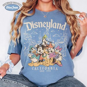 May include: Blue t-shirt with the word "Disneyland" and a graphic of Disney characters. The shirt also features the text "CALIFORNIA EST. 1955". The shirt is a Comfort Colors Blue Jean.