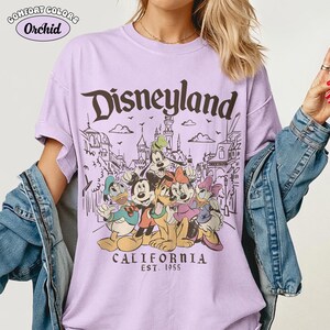 May include: A light purple t-shirt with the word "Disneyland" in brown script. The shirt features a graphic of Disney characters and the text "CALIFORNIA EST. 1955". The shirt is a Comfort Colors brand.