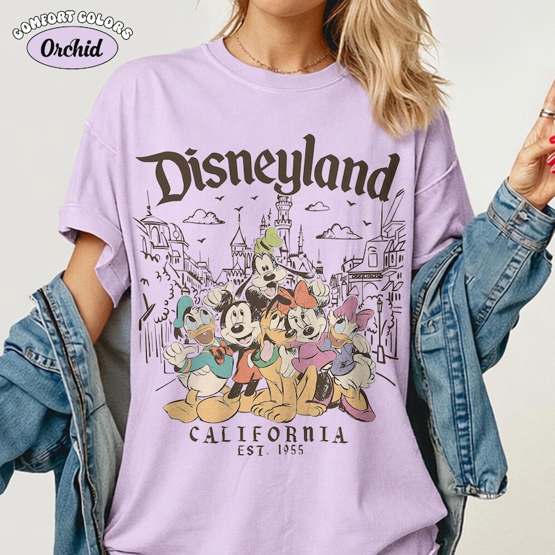 May include: A light purple t-shirt with the word "Disneyland" in brown script. The shirt features a graphic of Disney characters and the text "CALIFORNIA EST. 1955". The shirt is a Comfort Colors brand.