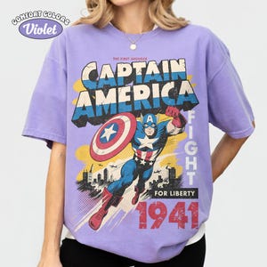May include: A violet short-sleeve t-shirt featuring a graphic print of Captain America with a shield, city skyline, and the text "The First Avenger Captain America Fight For Liberty 1941". The shirt has a Comfort Colors Violet label.