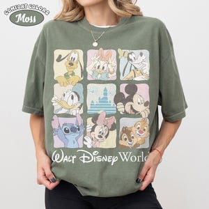 Op de afbeelding: Een mosgroen T-shirt met korte mouwen en een raster van negen vierkanten met illustraties van Disney-figuren en een kasteel. Onder het raster staat de tekst Walt Disney World in wit schrift. Het shirt heeft een ronde hals en een wijde pasvorm.