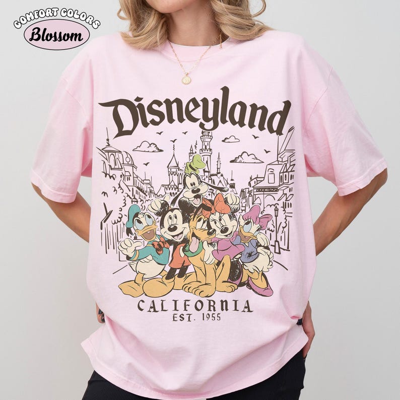 May include: Pink t-shirt featuring the word "Disneyland" in a brown, vintage font, with a graphic of Disney characters and a California theme. The shirt includes the text "CALIFORNIA EST. 1955".