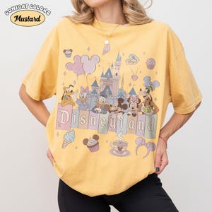 May include: Mustard-colored t-shirt with a Disneyland graphic. The graphic features Disney characters, a castle, and the word "Disneyland" in pastel colors. The shirt has short sleeves and a relaxed fit.