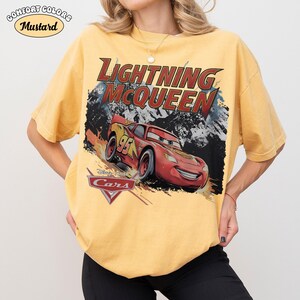 May include: A mustard yellow short-sleeve t-shirt featuring a graphic print of the red race car Lightning McQueen with the number 95. The text reads Lightning McQueen and Disney Pixar Cars. The shirt has a crew neckline and a relaxed fit.