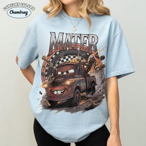 May include: Light blue Comfort Colors t-shirt featuring a graphic of Mater from the Cars movie. The design includes the word "MATER" in large letters above the character, with a racing theme background. The shirt is a short-sleeved crew neck.