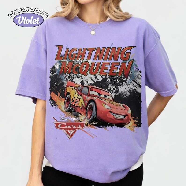 May include: A light purple short-sleeve t-shirt featuring a graphic of the red race car Lightning McQueen from the movie Cars. The text reads Lightning McQueen and Disney Pixar Cars. The shirt has a crew neckline and a relaxed fit design.