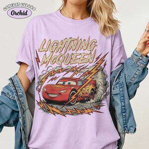 May include: A light purple Comfort Colors t-shirt featuring a graphic of Lightning McQueen from the Cars movie. The design includes the text "LIGHTNING McQUEEN" and a red race car with the number 95, along with lightning bolts.