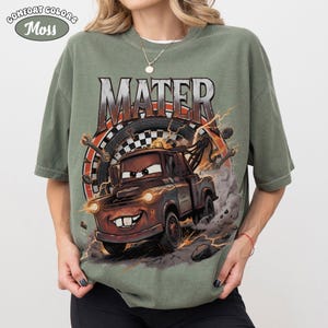 May include: Green Comfort Colors t-shirt featuring a graphic of Mater from the movie Cars. The design includes the word "MATER" above the cartoon tow truck, with lightning bolts and a tire in the background. The shirt has short sleeves.