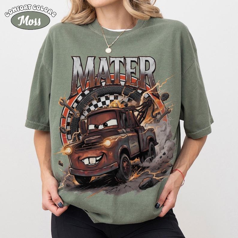 May include: Green Comfort Colors t-shirt featuring a graphic of Mater from the movie Cars. The design includes the word "MATER" above the cartoon tow truck, with lightning bolts and a tire in the background. The shirt has short sleeves.