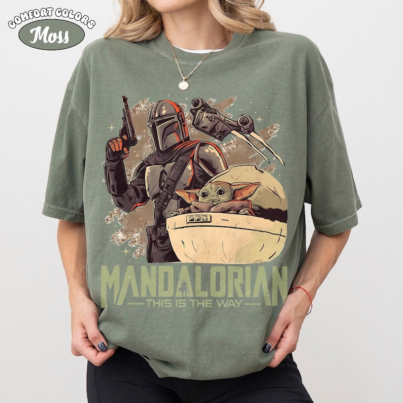 May include: A moss green t-shirt featuring a graphic print of a Mandalorian character holding a blaster, a small creature in a floating pod, and a starship. The text below the graphic reads MANDALORIAN THIS IS THE WAY in large letters.