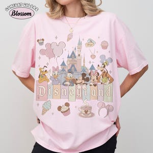 May include: A light pink t-shirt featuring a Disneyland graphic. The graphic includes cartoon characters, a castle, balloons, and the word "Disneyland". The shirt also has the text "Comfort Colors Blossom" in a circle.
