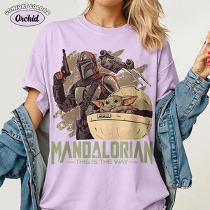 May include: A light purple t-shirt featuring a graphic print of a Mandalorian character holding a blaster, a small creature in a floating pod, and a starship. The text "MANDALORIAN" and "THIS IS THE WAY" is printed in green below the graphic.
