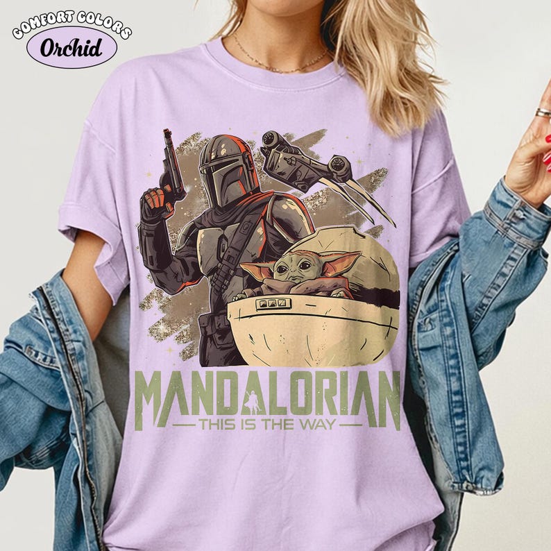 May include: A light purple t-shirt featuring a graphic print of a Mandalorian character holding a blaster, a small creature in a floating pod, and a starship. The text "MANDALORIAN" and "THIS IS THE WAY" is printed in green below the graphic.