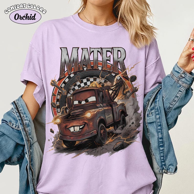 May include: A light purple Comfort Colors t-shirt featuring a graphic of Mater from the Cars movie. The design includes the word "MATER" in a distressed font, a racing wheel, and lightning bolts. The shirt has short sleeves.