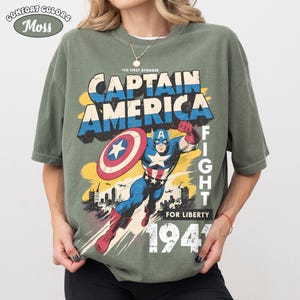 May include: A moss green t-shirt featuring a graphic print of Captain America in a dynamic pose with a shield. The text reads THE FIRST AVENGER, CAPTAIN AMERICA, FIGHT FOR LIBERTY, and 1941. The design includes a city skyline and airplanes.