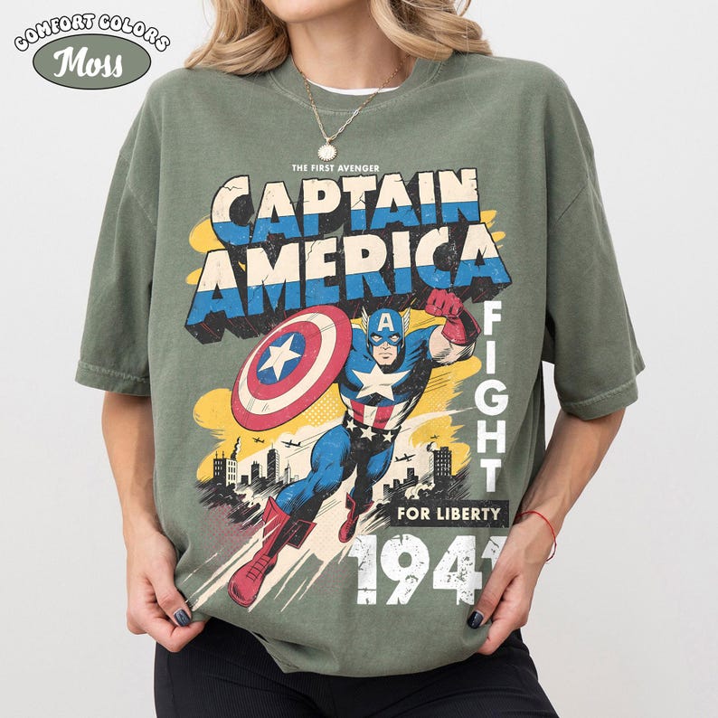 May include: A moss green t-shirt featuring a graphic print of Captain America in a dynamic pose with a shield. The text reads THE FIRST AVENGER, CAPTAIN AMERICA, FIGHT FOR LIBERTY, and 1941. The design includes a city skyline and airplanes.