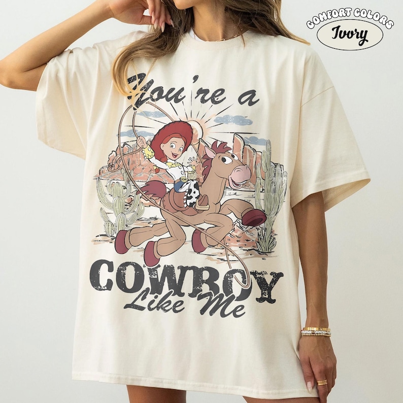 May include: An ivory t-shirt featuring a graphic of a cartoon cowgirl riding a brown horse with a lasso. The text reads "You're a COWBOY Like Me" above and below the illustration, with a desert landscape background including cacti and mountains.