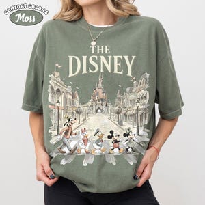 May include: Olive green t-shirt with the text "THE DISNEY" above a graphic of Disney characters crossing a street in front of a castle. The shirt has the words "COMFORT COLORS" and "Moss" in a circle at the top left.
