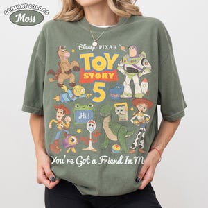 May include: A moss green Comfort Colors t-shirt featuring a colorful graphic of Toy Story characters. The design includes Woody, Buzz Lightyear, and other characters from the movie, with the text "TOY STORY 5" and "You're Got a Friend In Me".