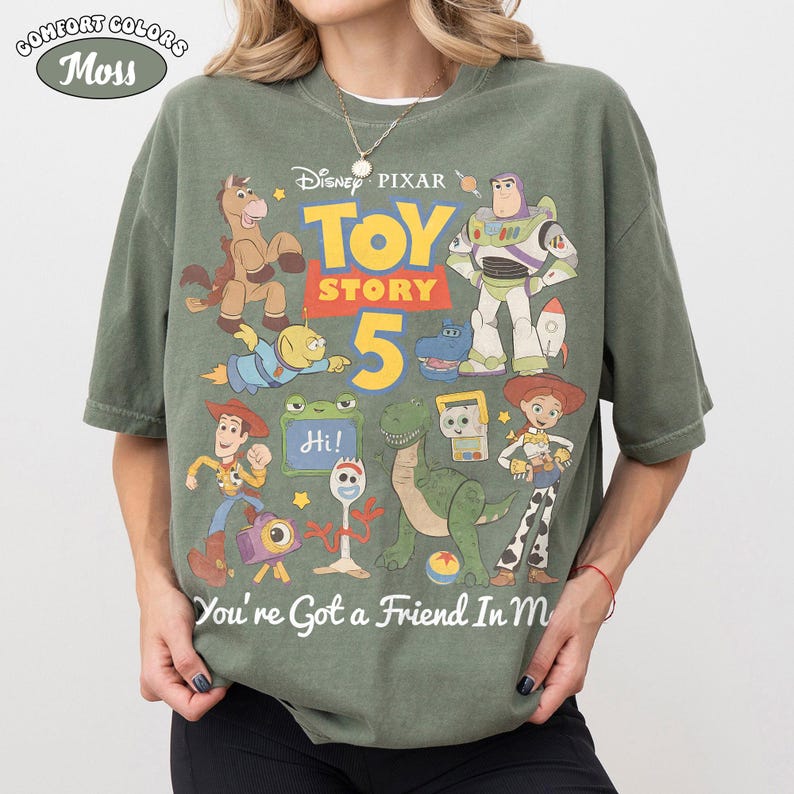 May include: A moss green Comfort Colors t-shirt featuring a colorful graphic of Toy Story characters. The design includes Woody, Buzz Lightyear, and other characters from the movie, with the text "TOY STORY 5" and "You're Got a Friend In Me".
