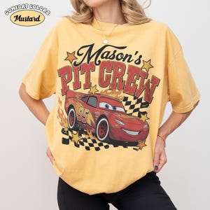 May include: Mustard-colored t-shirt with a graphic of Lightning McQueen from the movie Cars. The shirt features the text "Mason's Pit Crew" and the number "95". The shirt has a vintage look.