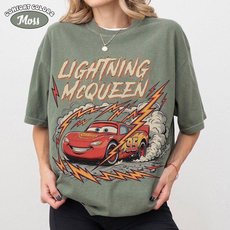 May include: Olive green t-shirt featuring Lightning McQueen from the Cars movie. The design includes the text "Lightning McQueen" in a vintage font, along with a graphic of the red race car and lightning bolts.