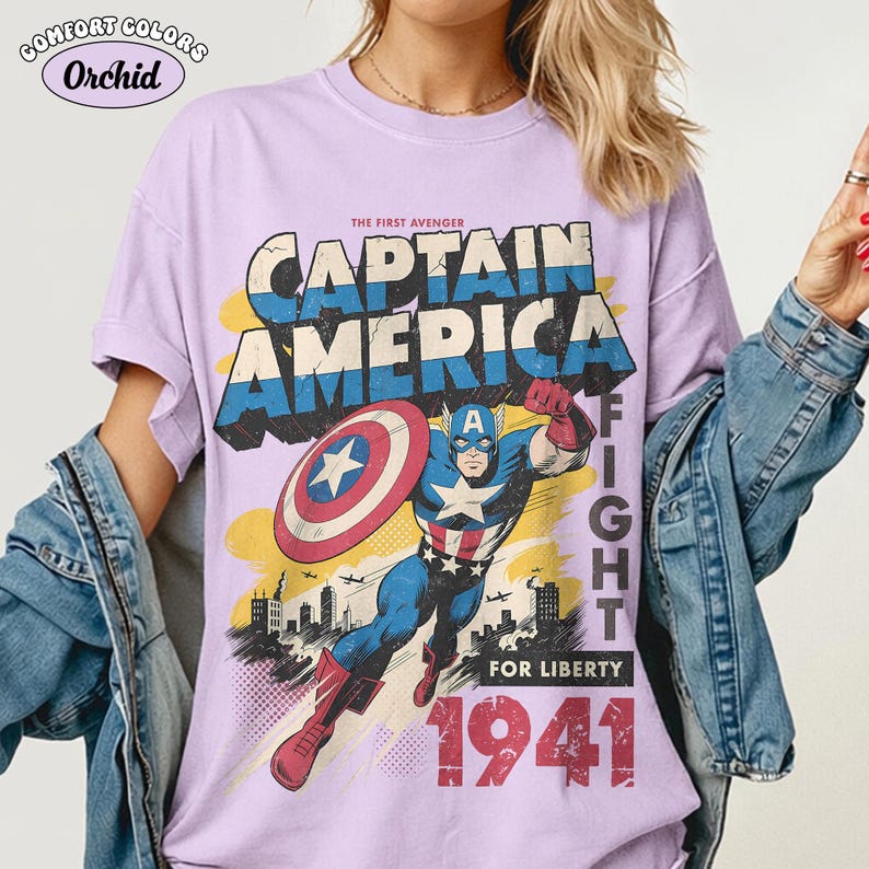 May include: A light purple t-shirt featuring a vintage-style graphic of Captain America holding a shield. The text reads THE FIRST AVENGER CAPTAIN AMERICA FIGHT FOR LIBERTY 1941. The design includes a city skyline, airplanes, and a yellow background.