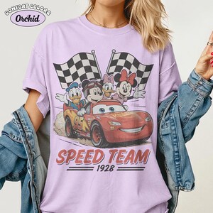 May include: A lilac t-shirt featuring a graphic of Disney characters in a red race car. The car has the number 95 on it. The shirt also has the words "SPEED TEAM 1928" printed on it. The shirt has the words "COMFORT COLORS Orchid" printed on it.