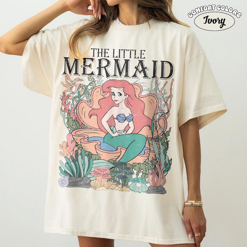 May include: Ivory-colored t-shirt featuring a vintage-style graphic of Ariel from "The Little Mermaid." The design includes Ariel in a seashell, surrounded by aquatic plants. The text "THE LITTLE MERMAID" is above the graphic. The shirt is labeled "COMFORT COLORS" and "Ivory."