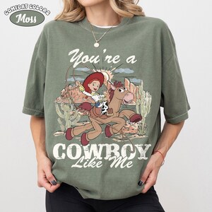 May include: A moss green short-sleeve t-shirt featuring a graphic print of a cowgirl riding a horse with a lasso. The design includes a desert landscape with cacti and the text "You're a COWBOY Like Me" in a distressed white font style.