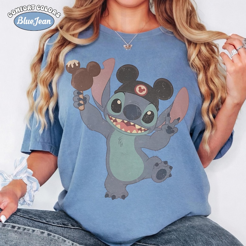 May include: A blue short-sleeve t-shirt featuring a graphic of the character Stitch wearing a black hat with mouse ears and holding a chocolate-covered treat. The shirt includes a Comfort Colors Blue Jean logo in the upper left corner.