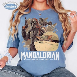 May include: A light blue t-shirt featuring a graphic of a Mandalorian character holding a blaster, a small green creature in a floating pod, and a starship. The text "MANDALORIAN" and "THIS IS THE WAY" is printed below the graphic in white lettering.
