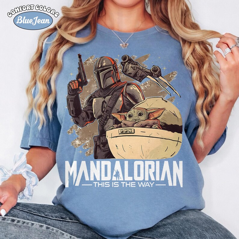 May include: A light blue t-shirt featuring a graphic of a Mandalorian character holding a blaster, a small green creature in a floating pod, and a starship. The text "MANDALORIAN" and "THIS IS THE WAY" is printed below the graphic in white lettering.