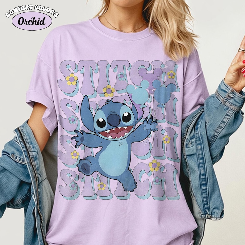May include: A light purple short-sleeve t-shirt featuring a graphic of the blue character Stitch holding two balloons. The text STITCH is repeated in a wavy font behind the character, accented with small yellow and blue flowers and Mickey icons.