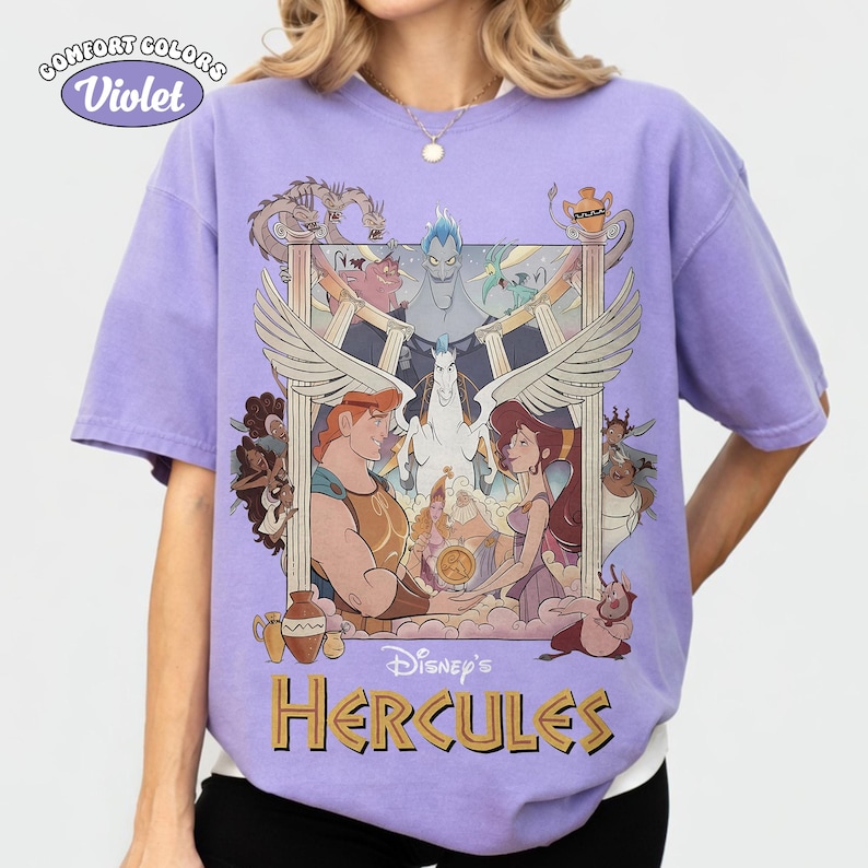 May include: Violet Comfort Colors t-shirt featuring a graphic from Disney's Hercules. The design includes Hercules, Megara, Pegasus, and other characters. The text "Disney's Hercules" is printed in gold. The shirt has short sleeves.