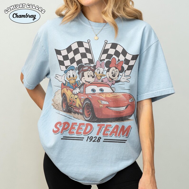 May include: Pale blue t-shirt with a graphic of Mickey Mouse, Donald Duck, Daisy Duck, and Minnie Mouse in a red race car. The text "SPEED TEAM 1928" is below the image. The shirt has the Comfort Colors Chambray logo.