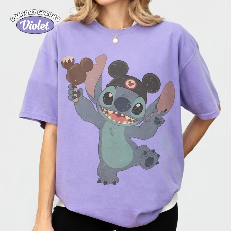 May include: A violet short-sleeve t-shirt featuring a graphic of the character Stitch wearing black mouse ears and holding a brown chocolate-covered treat. The text Comfort Colors Violet is printed in the upper left corner of the garment.