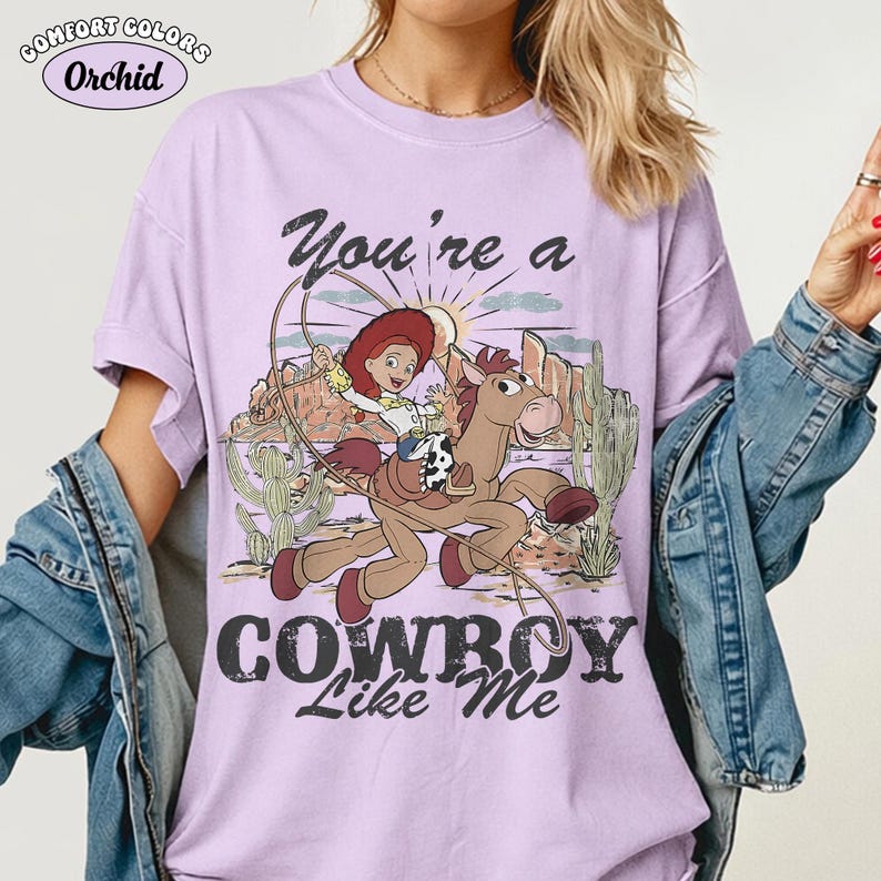 May include: A light purple short-sleeve t-shirt featuring a graphic of a cowgirl riding a horse in a desert landscape with cacti and mountains. The text reads You're a COWBOY Like Me. A Comfort Colors Orchid label is visible in the top left corner.