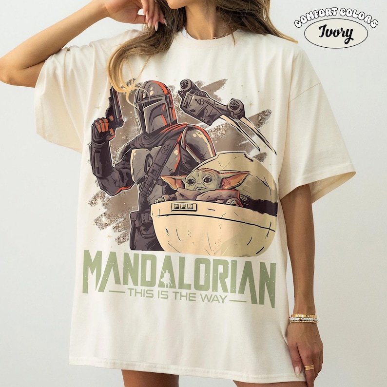May include: An ivory t-shirt featuring a graphic print of a Mandalorian character holding a blaster, a small creature in a floating pod, and a starship. The text reads "MANDALORIAN" and "THIS IS THE WAY" in green, with a "COMFORT COLORS IVORY" logo.