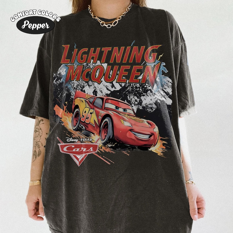 May include: A charcoal gray short-sleeve t-shirt featuring a graphic of the character Lightning McQueen from the Disney Pixar movie Cars. The design includes the text Lightning McQueen and the Cars logo, with a distressed, faded aesthetic.