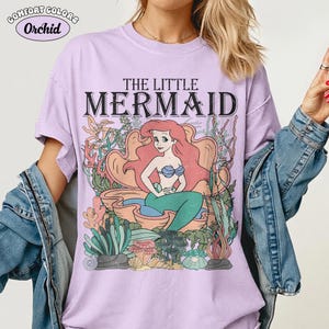 May include: A lilac Comfort Colors t-shirt featuring a graphic of Ariel from The Little Mermaid. The design includes the text "THE LITTLE MERMAID" above an illustration of Ariel in a seashell, surrounded by coral and seaweed.