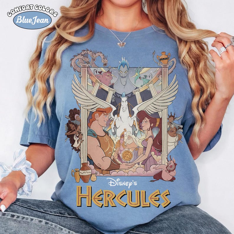 May include: Blue t-shirt with a graphic from Disney's Hercules. The design features Hercules, Megara, Pegasus, and other characters in a framed illustration. The shirt has the text "Disney's Hercules" in gold letters and the words "Comfort Colors Blue Jean" in a circle.