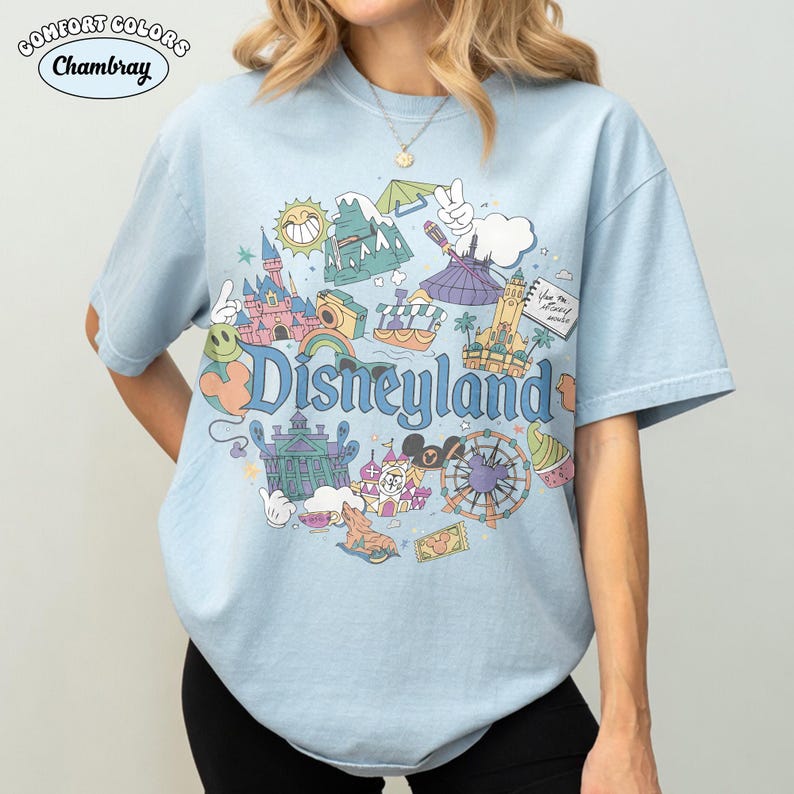 May include: Pale blue t-shirt featuring a Disneyland graphic with various park icons. The design includes a castle, Matterhorn, and a Ferris wheel. The shirt has the words "Comfort Colors" and "Chambray" printed on it.
