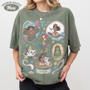 May include: Moss green Comfort Colors t-shirt featuring a colorful graphic of characters from the animated film Moana. The design includes images of Maui, Moana, Pua, Hei Hei, and other elements from the movie.