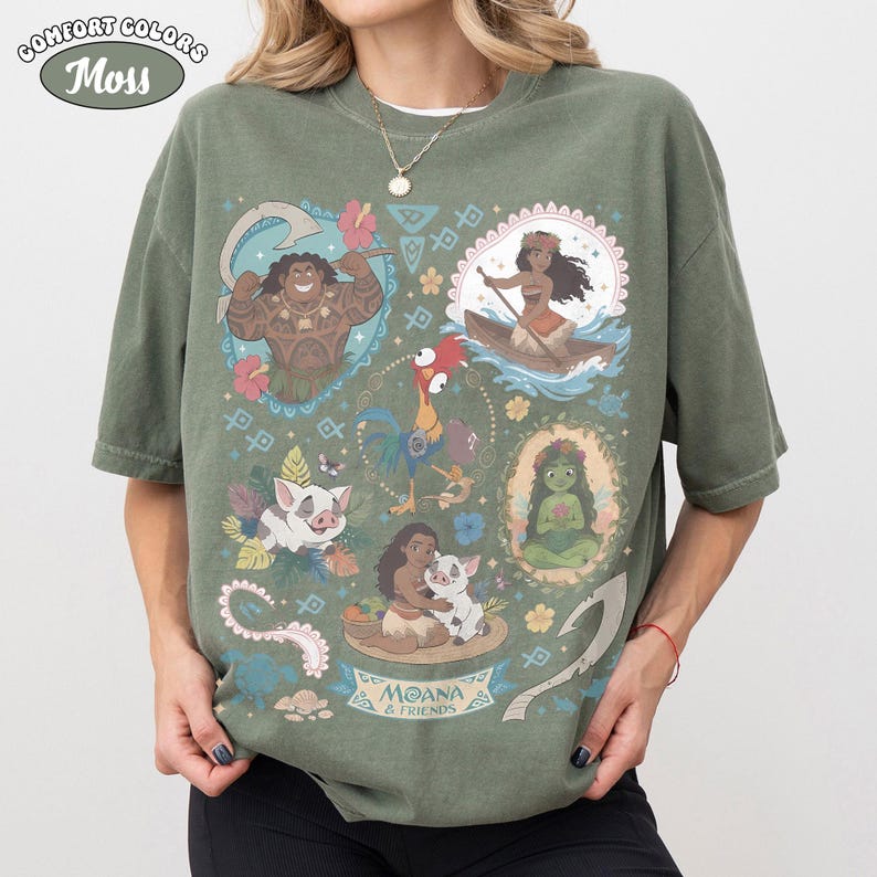 May include: Moss green Comfort Colors t-shirt featuring a colorful graphic of characters from the animated film Moana. The design includes images of Maui, Moana, Pua, Hei Hei, and other elements from the movie.