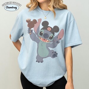 May include: A light blue short-sleeve t-shirt featuring a graphic of the character Stitch wearing black mouse ears and holding a brown chocolate treat. The text Comfort Colors and Chambray appears in the upper left corner of the image.